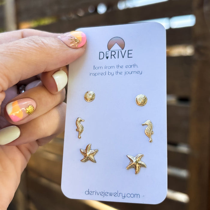 Gold sea-themed earrings on a Derive jewelry card held by a hand with colorful nail polish.