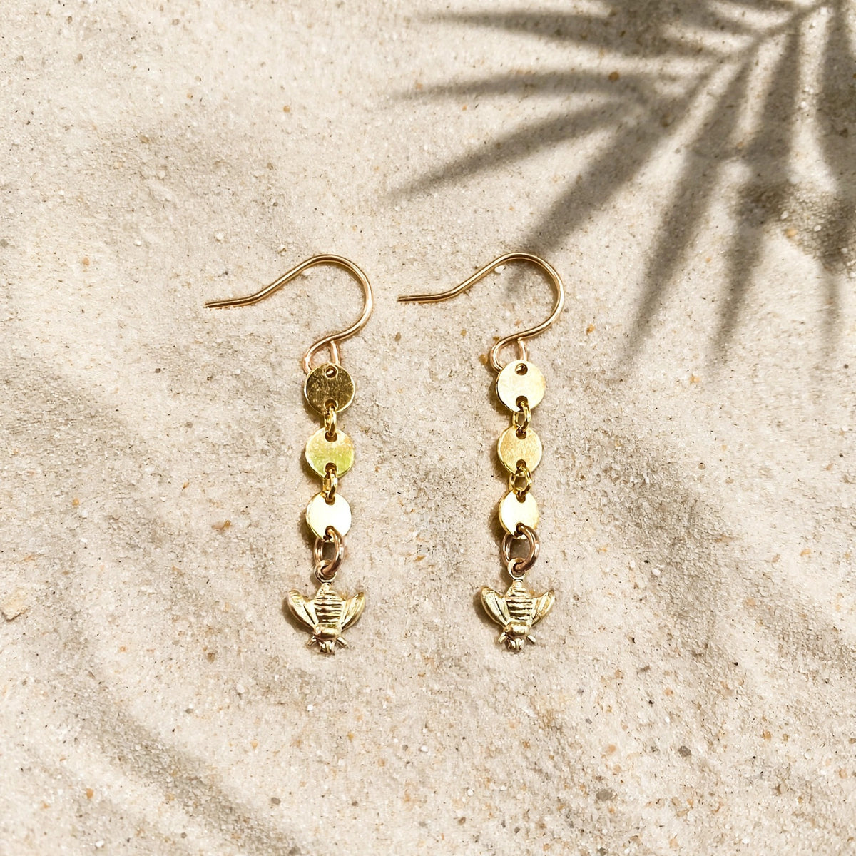 Bee Charm Coin Earrings in 14k gold fill, styled on pale sand at the beach