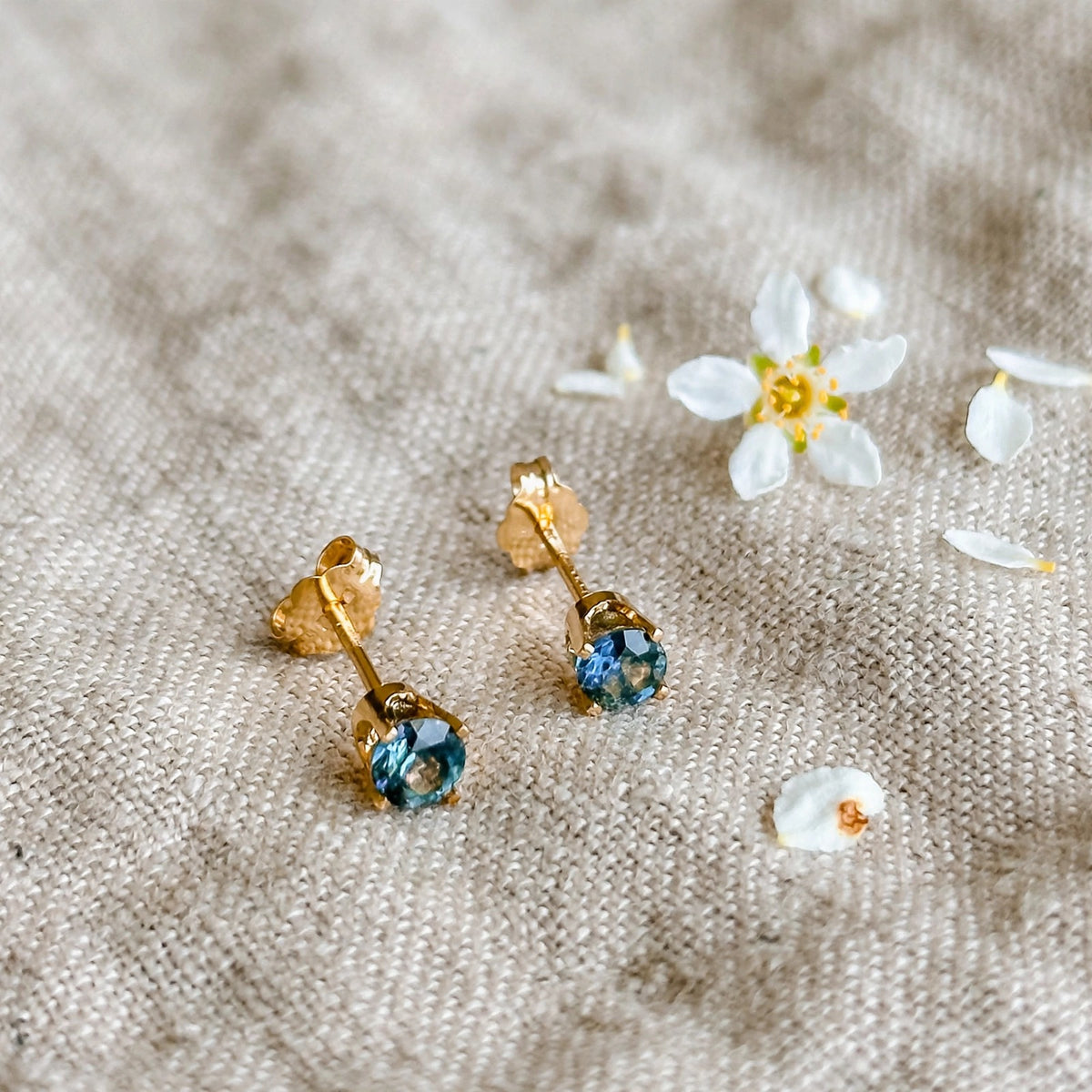 Montana sapphire stud earrings in solid 14k gold beside a white blossom on natural linen