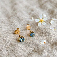 Montana sapphire stud earrings in solid 14k gold beside a white blossom on natural linen
