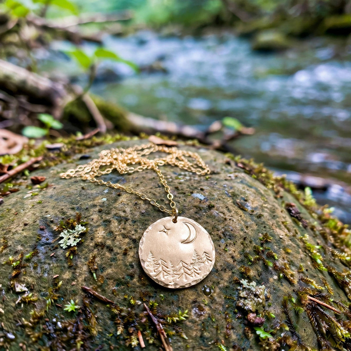 Moonlit Forest Hand Stamped Coin Necklace in 14k gold fill with a moon, star and pine tree coin pendant, resting on a rock by a forest stream