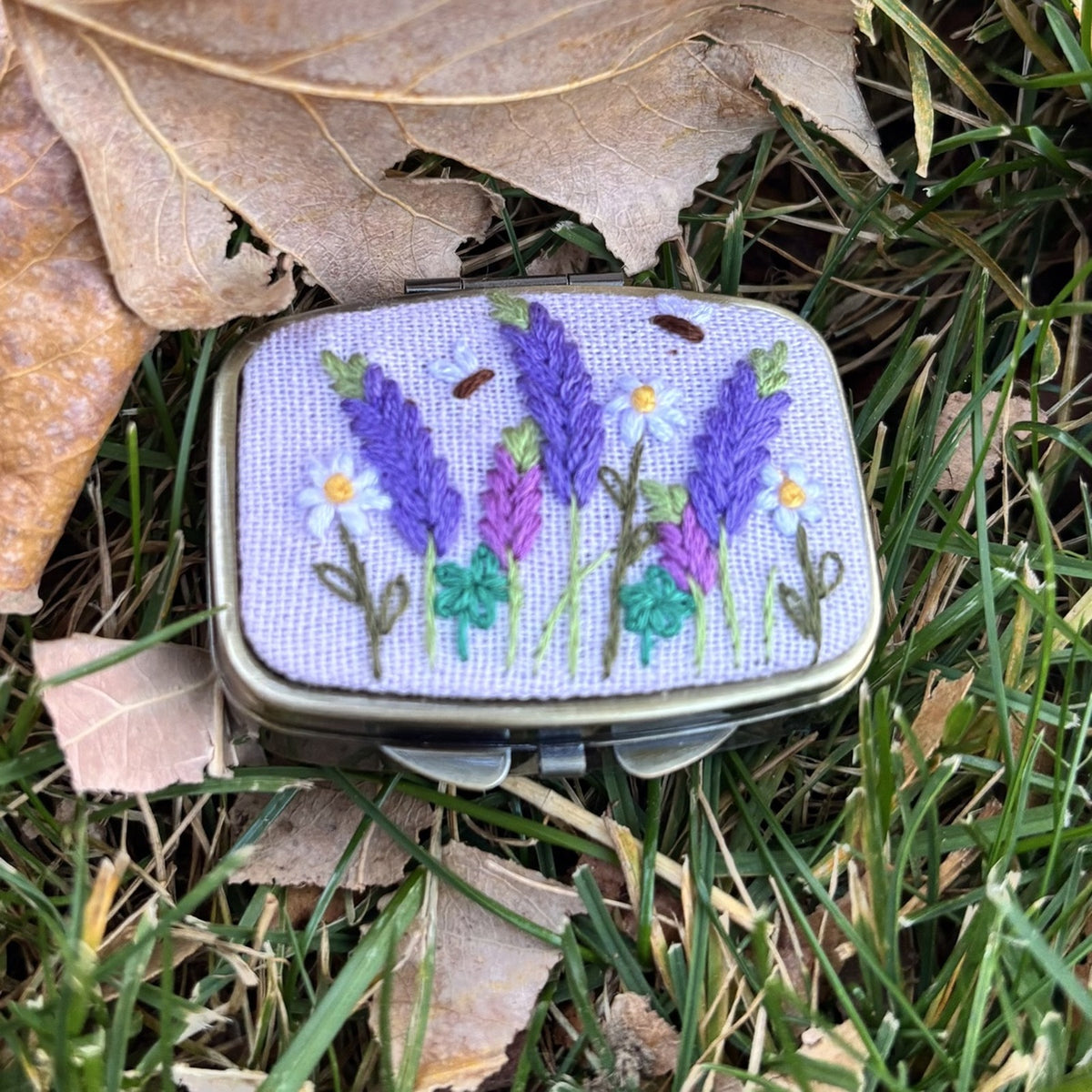 Rectangular metal jewelry case in light lavender with lavender sprig and daisy embroidery, front view on grass