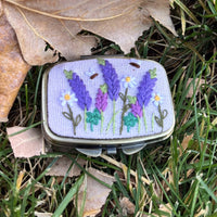 Rectangular metal jewelry case in light lavender with lavender sprig and daisy embroidery, front view on grass