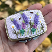 Rectangular metal jewelry case in light lavender with lavender sprig and daisy embroidery, front view held in hand