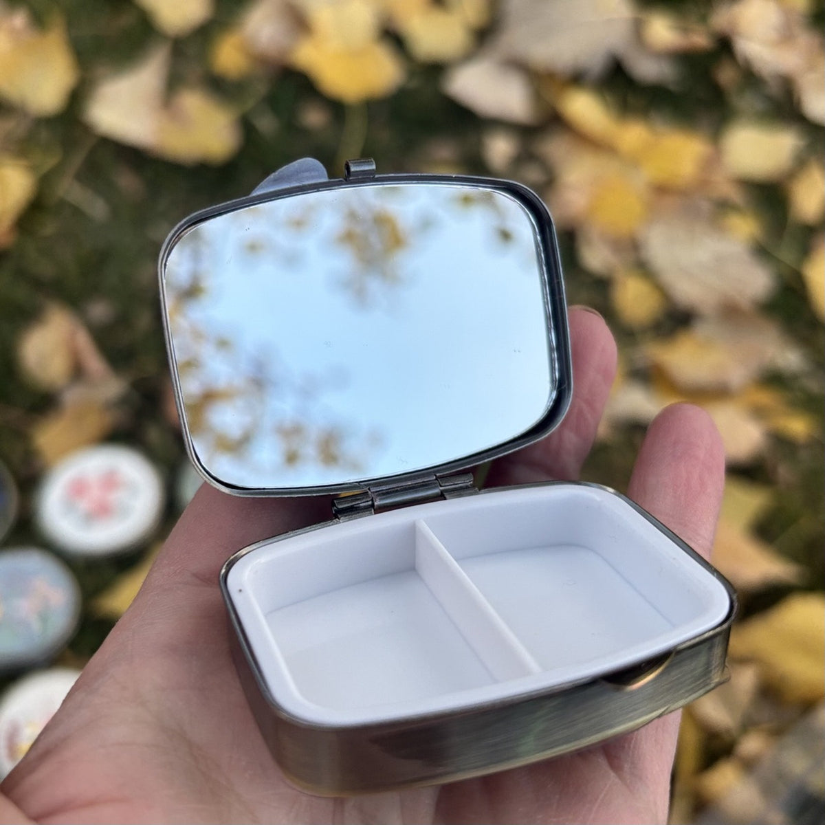 Rectangular metal jewelry case in light lavender, open in hand showing the mirror and two compartment tray