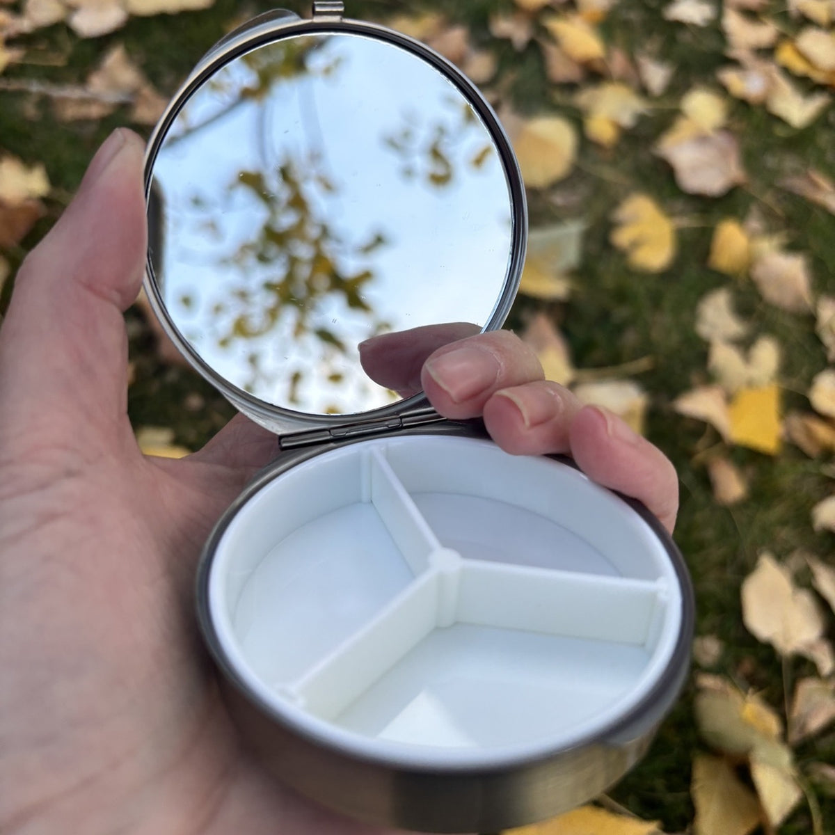 Round metal jewelry case with blue grey embroidered lid, open in hand showing the mirror and three compartment tray