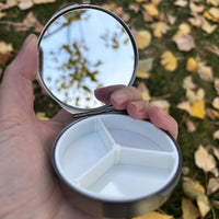 Round metal jewelry case with blue grey embroidered lid, open in hand showing the mirror and three compartment tray