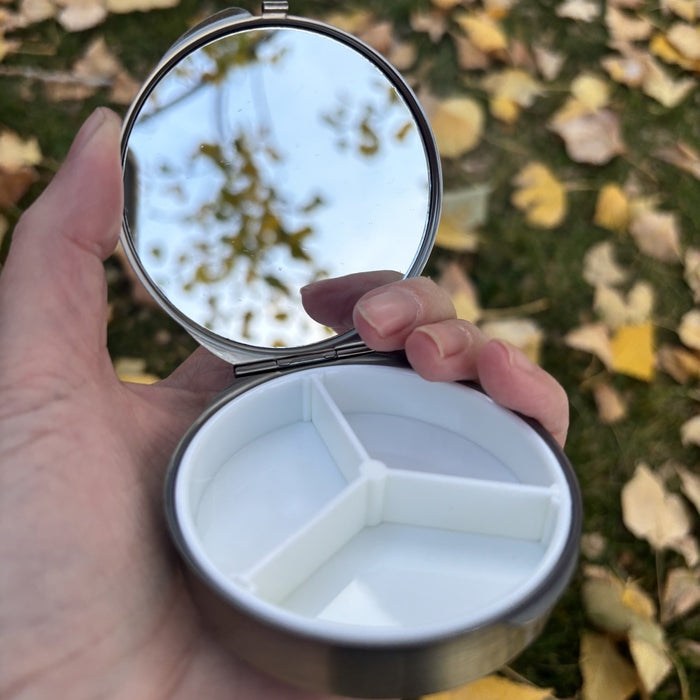 Round metal jewelry case with blue grey embroidered lid, open in hand showing the mirror and three compartment tray