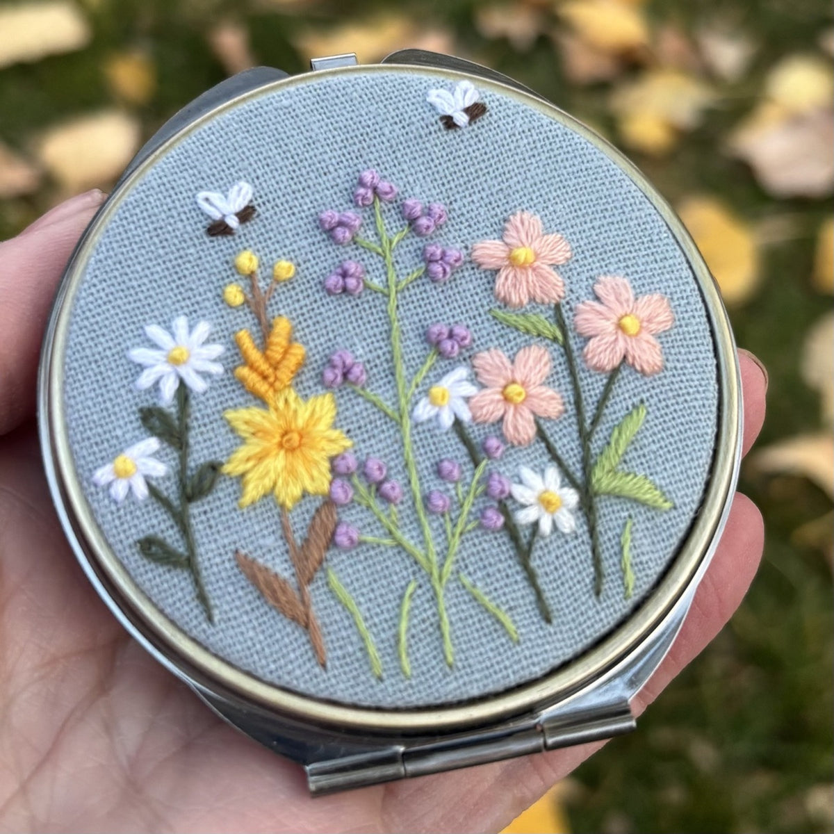 Round metal jewelry case in blue grey with wildflower and butterfly embroidery, closed front view held in hand