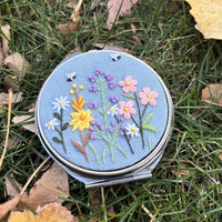 Round metal jewelry case in blue grey with wildflower and butterfly embroidery, closed front view on grass