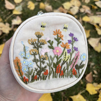 Round embroidered floral travel jewelry case in off white with wildflower and bee embroidery, front view held in hand