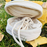 Round embroidered travel jewelry case in off white, open on grass showing the drawstring pouch inside