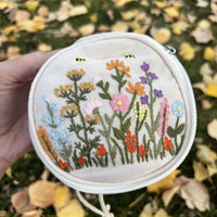 Round embroidered floral travel jewelry case in off white with wildflower and bee embroidery, front view showing the zipper