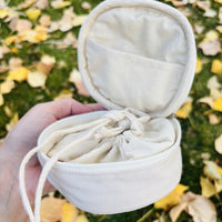 Round embroidered travel jewelry case in off white, open in hand showing the lid pocket and drawstring pouch inside