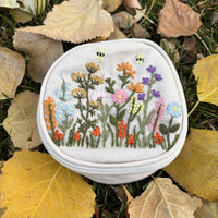 Round embroidered floral travel jewelry case in off white with wildflower and bee embroidery, front view on autumn leaves