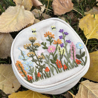 Round embroidered floral travel jewelry case in off white with wildflower and bee embroidery, angled view on autumn leaves showing the zipper