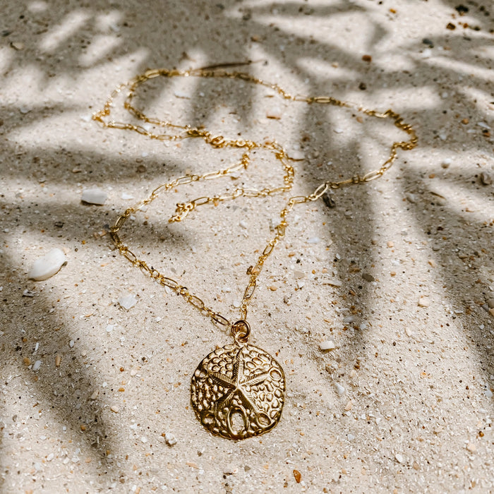 Sand Dollar Pendant Necklace in 14k gold fill, styled on pale sand at the beach