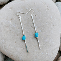 Sticks and Stones Kingman Turquoise Stick Earrings in sterling silver, styled on pale sand at the beach