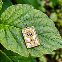 The sun tarot card pendant in solid 14k gold resting on a fresh green leaf